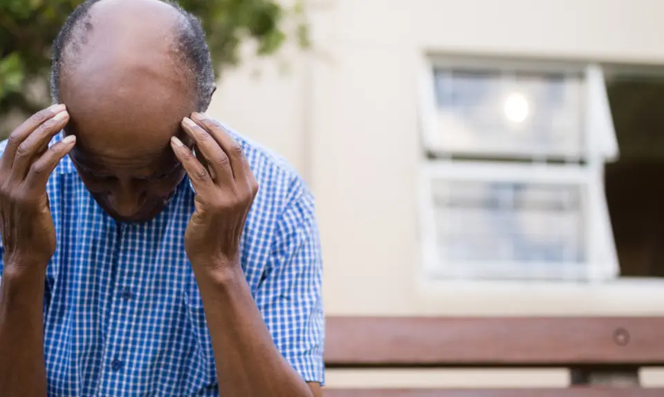 An elderly man sits on a bench and holds his head in his hands.