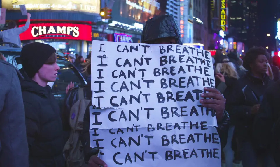 A man holds a sign in a crowded city; the sign says "I can't breathe. I can't breathe. I can't breathe."