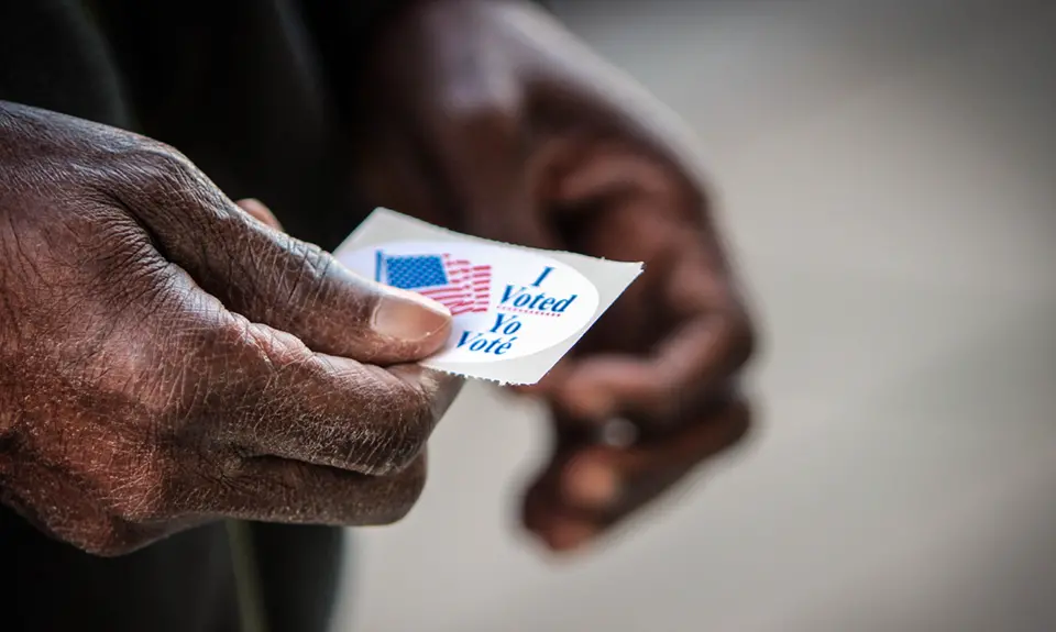 A close up of an elderly Black man's hands. He has just finished casting his ballot, and is holding his “I voted” sticker.