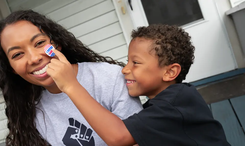 A Black son places an "I Voted" sticker on his mother's smiling face.