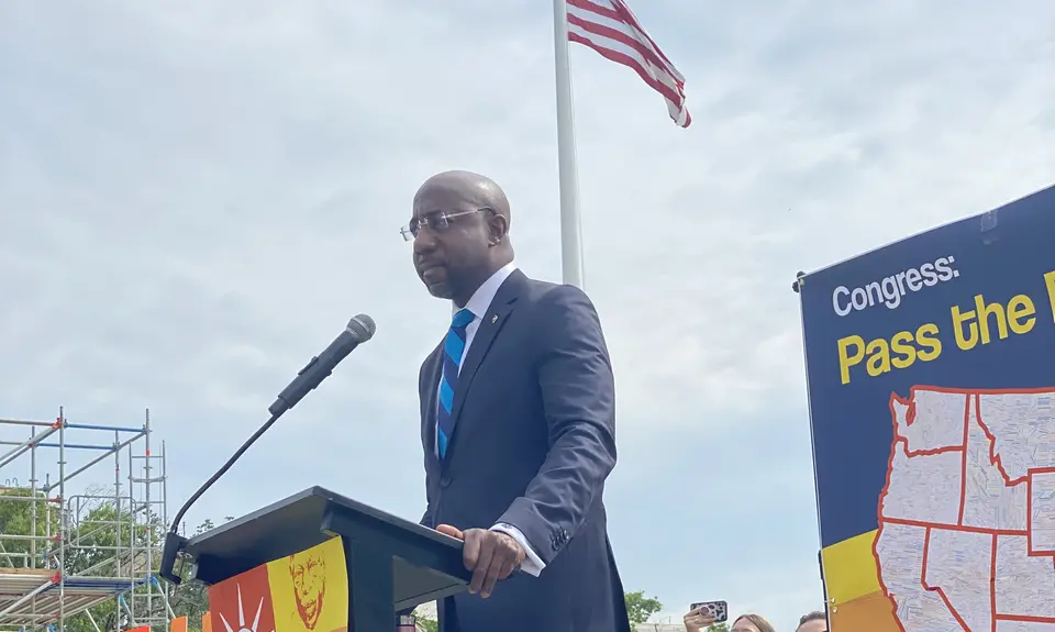 Sen. Raphael Warnock standing at a podium outdoors with an American flag in the background.