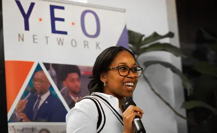 Woman speaking into a microphone in front of a sign that says YEO Network