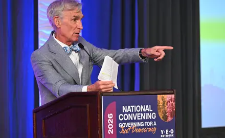 Bill Nye wearing a gray suit and blue bow tie standing at a podium at the Young Elected Officials Network Convening 2026 and holding a copy of the Constitution. 