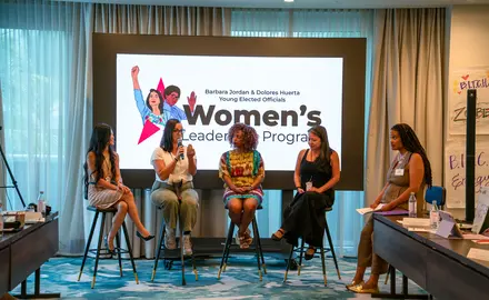Five women sit on stools in front of a sign that reads "Women's Leadership Program"