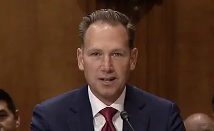Brian Burch, a white man with receding hairline wearing blue suit and red tie seated at a table at his confirmation hearing.