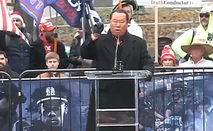 Che Ahn speaking at microphone at outdoors Jan. 5, 2021 rally on Freedom Plaza near the White House. Ahn is wearing black coat and gloves, people holding Trump flags are visible behind him.