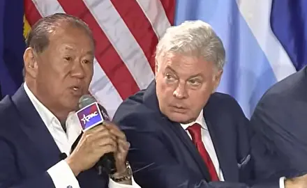 Che Ahn speaks into a microphone branded with a CPAC logo; seated to his left and listening to Ahn is Lance Wallnau.