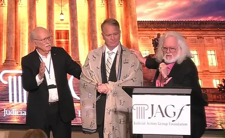 Dominionist Chuck Pierce, who has a gray beard and long gray hair, stands behind a lectern with the logo for the Judicial Action Group Foundation logo. His right hand rests on the shoulder of Phillip Jauregui, who has a shawl embroidered with crowns draped over his shoulders. A third man has his hand up in prayer. They are standing before an image of the U.S. Supreme Court building lit up at night. 