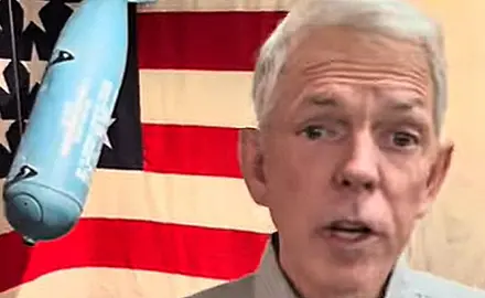 David Barton speaks into the camera; visible behind him is a World War II era (?) bomb hanging in front of a U.S. flag.