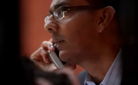 Close up side-angle view of Dinesh D'Souza looking serious and looking off camera to the left while holding a cell phone to his right ear.