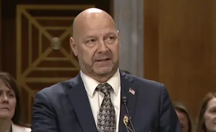 Doug Mastriano in suit and tie is seated at a table in a Senate hearing room, speaking to an off-camera senator