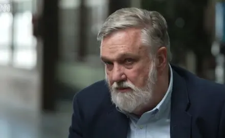 Douglas Wilson, a white man with gray hair and beard, is looking off camera to the CNN reporter interviewing him. The background is blurred. 