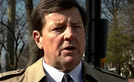 Ed Martin, wearing suit and tie and tan overcoat, is being interviewed outdoors; visible behind him are neighborhood homes and trees. 