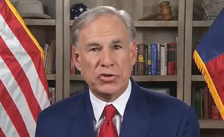 Gov. Greg Abbott wearing blue suit and red tie speaks to the camera; bookshelves are visible behind him with an American flag partially visible to the left and Texas flag to the right