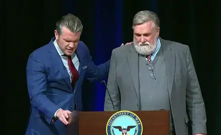 Defense Secretary Pete Hegseth and Christian nationalist pastor Doug Wilson praying at lectern bearing the logo of the "Department of War." Their heads are bowed, and Hegseth has one hand on the lectern and one on Wilson's shoulder.