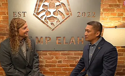 MAGA musician Sean Feucht and then-Senate candidate Hung Cao are seated in front of a brick wall on which is huge a large sign identifying their location as "Camp Elah," the Capitol Hill outpost for Feucht's political activities.