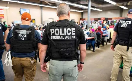 Photo shows several officers identified as police and/or ICE from the back as they face workers gathered in what appears to be a workplace lunchroom