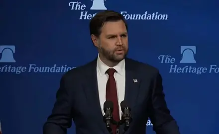 J.D. Vance, wearing suit and tie, stands behind lectern with Heritage Foundation logo behind him.