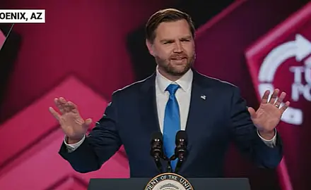 Vice President J.D. Vance gestures with his hands while speaking at lectern on stage at TPUSA's AmericaFest; the TPUSA logo is partially visible behind him.