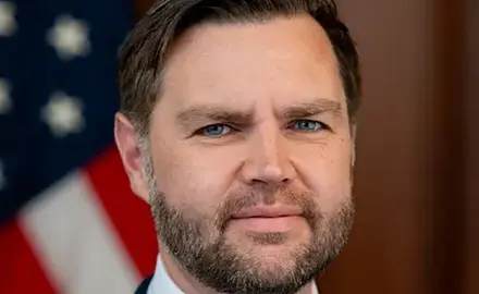 Close-up photo portrait of J.D. Vance with a blurry US flag visible behind him 