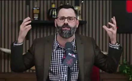 Joel Webbon sitting at a microphone in his studio, both hands raised in the air as he makes a point
