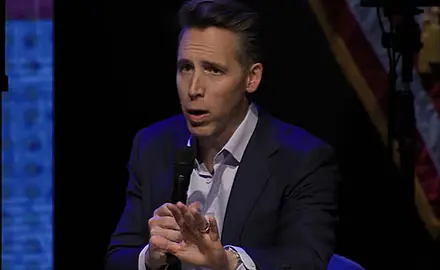 Sen Josh Hawley holds a microphone in his right had and gestures toward the audience with his left hand while seated on stage