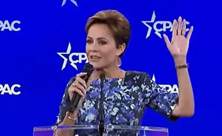 Kari Lake on stage in front of CPAC logos; she holds microphone in her right hand with her left hand raised in the air. 