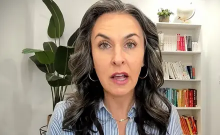 Katy Faust, a white woman with long black hair streaked with gray, speaks into the camera; a bookshelf and plant are visible behind her
