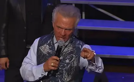 Kent Christmas, standing on stage at the Ryman Auditorium, speaks into a microphone in his right hand while pointing toward the crowd with his left hand; he looks angry.