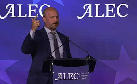 Heritage Foundation President Kevin Roberts, a bald white man wearing suit and tie, gestures with his right hand while speaking behind a lectern bearing the ALEC logo, which is also repeated on the background. 
