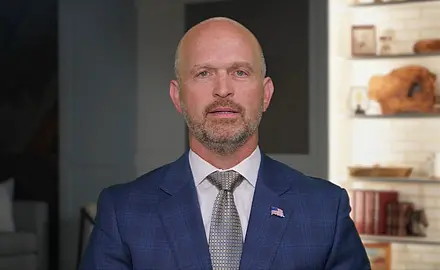 Heritage Foundation President Kevin Roberts, in suit and tie, speaks into a camera standing in what looks like an office, with a bookshelf behind him.