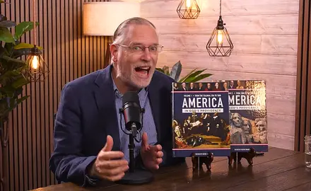 Kevin Swanson speaks animatedly into a microphone; two copies of his 1,000 page textbook on America's "Christian history" are propped up on the table next to him. 