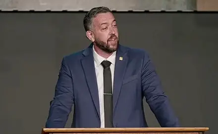 Kyle Lovett, a bearded white man in suit and tie, speaks into a mic in front of a nondescript background at an event at his Wichita, Kansas, church