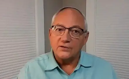 Glenn Clary, a balding white man with gray hair and wearing glasses, is seated in a nondescript room wearing a light blue polo shirt.