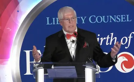Mat Staver, a white man wearing a suit and red bowtie, gestures while speaking at lectern in front of logo for Liberty Counsel's 2025 gala and its theme "A New Birth of Freedom"