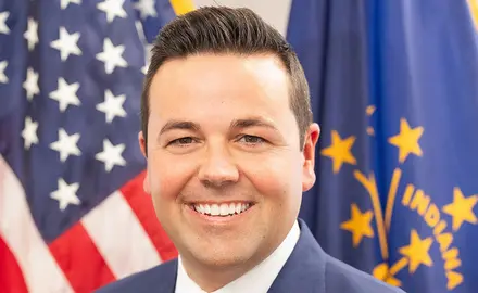 Portrait-style official photo of Indiana Lt. Gov. Micah Beckwith smiling into the camera with U.S. and Indiana flags visible behind him.