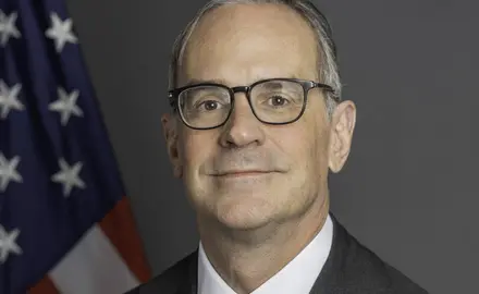 Formal portrait photo of Michael Anton in suit and tie looking into the camera with an American flag behind his right shoulder