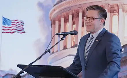 Image shows Mike Johnson standing and a lectern and speaking into a microphone in front of an illustrated image showing the Capitol dome and a U.S. flag fluttering in the breeze.