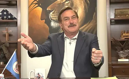 Mike Lindell, seated in front of a painting of a lion's head superimposed on the head of Christ, gestures with both hands. A cross and a US flag are visible on the shelves behind him. 