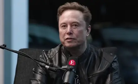 Elon Musk, in black shirt and leather jacket, is seated at a microphone that is emblazoned with a capital letter E, the logo for The Economist magazine