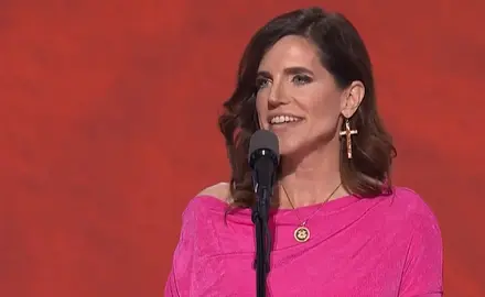 Rep. Nancy Mace, wearing bright pink dress and oversize cross-shaped earrings, speaks into a microphone in front of a red background on stage at the 2024 Republican National Convention.