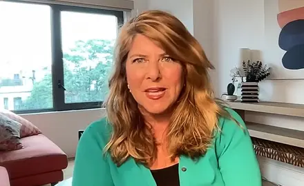 Naomi Wolf is seated in what appears to be a home office, with a window, shelf, and art visible behind her.