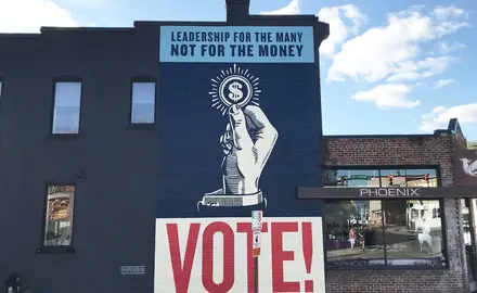 A mural with a handle holding a coin and text that reads "Leadership for the many, not for the money" and "Vote!"