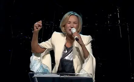 Paula White raises her right hand in a fist while speaking into a microphone held in her left hand. She is wearing a fancy white jacket; the background is dark. 