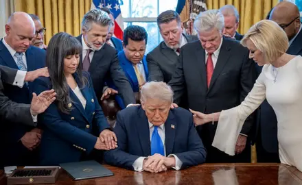 White House photo of Trump seated at his desk in the Oval Office surrounded by religious-right leaders; White House Faith Director Paula White, standing at right of image, lays her right hand on Trump's shoulder as she prays,