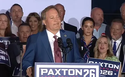 Texas Attorney General Ken Paxton stands at microphone behind a lectern with his Paxton 26 campaign logo. Supporters holding Paxton signs are gathered behind him.