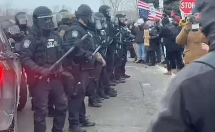 Image shows line of helmeted baton-wielding law enforcement officers facing off against protesters