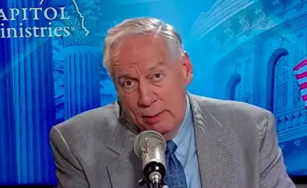 Ralph Drollinger, white man with gray hair in suite and tie, is seated and speaking into a microphone. The background includes the logo of Capitol Ministries and an illustrated collage of images including part of the Capitol dome and U.S. flag.