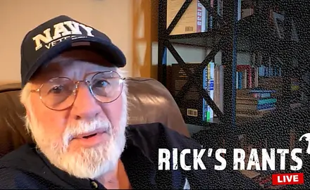 Rick Joyner, a white man with gray beard and mustache wearing a blue baseball cap with NAVY written on it, speaks into the camera while seated in an easy chair. 