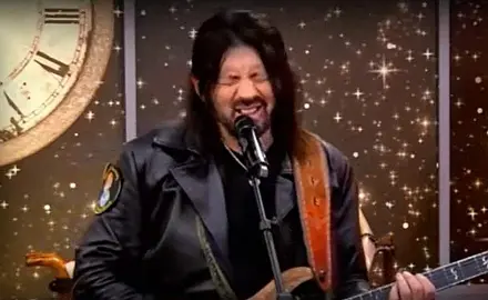 Robin Bullock, a white man with long black hair, grimace while speaking/singing into a microphone and holding a guitar. 